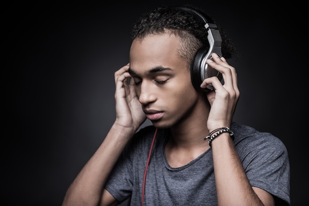 soul music. side view of young african man adjusting headphones and keeping eyes closed while standing against black background