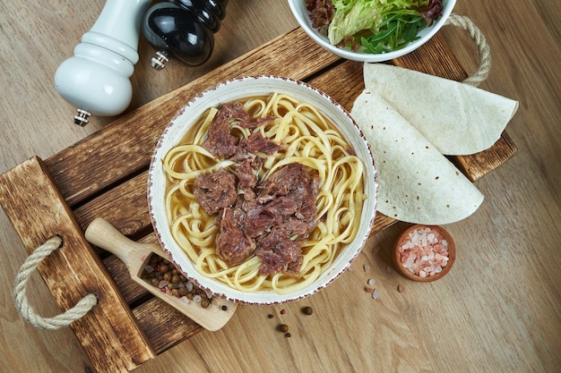 Soup on the broth with noodles and stewed beef in a white ceramic plate on a wooden tray. Food flat lay