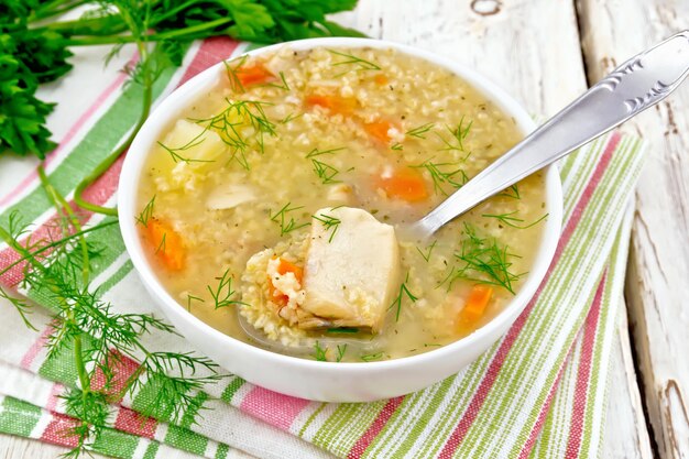 Zuppa di kulesh di pesce con miglio, patate, carote e cucchiaio in una ciotola bianca su un tovagliolo, prezzemolo, aneto su una tavola di legno chiara sullo sfondo