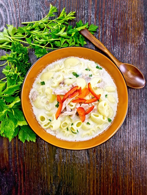 Soup from chicken meat, pasta with cream and sweet pepper, parsley and cilantro in a clay plate on a wooden board background from above