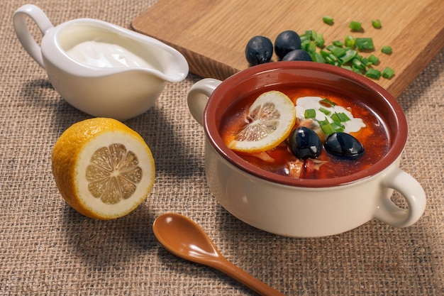 Soup saltwort with meat, smoked sausages, potatoes, tomatoes, marinated pickled cucumber, lemon, black olives and sour cream in ceramic soup bowl