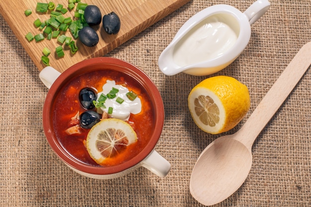 Soup saltwort with meat, smoked sausages, potatoes, tomatoes, marinated pickled cucumber, lemon, black olives and sour cream in white ceramic soup bowl with cutting board and wooden spoon