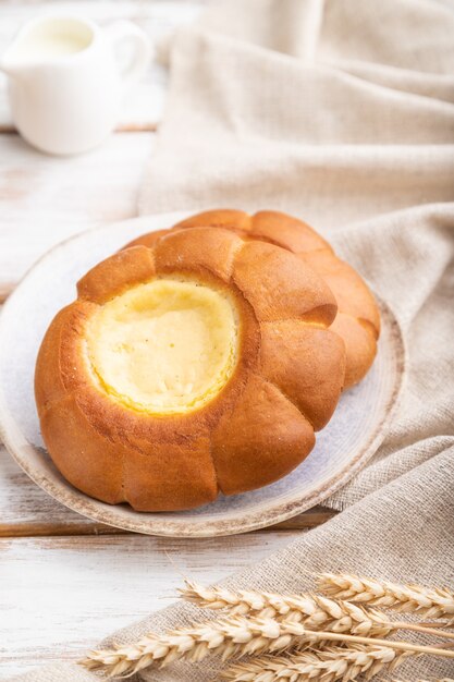 Sour cream bun with cup of coffee on a white wooden background and linen textile. Side view, close up, selective focus.