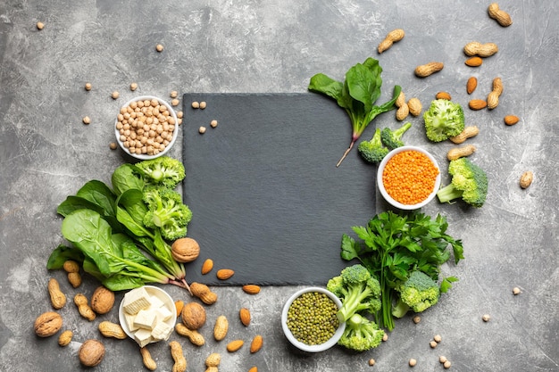 A source of protein for vegetarians. Healthy clean food: greens, vegetables, nuts and legumes top view on a concrete background with a black cutting stone in the center.