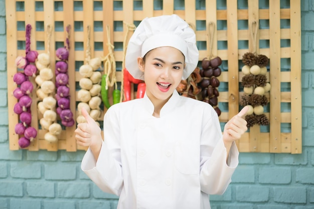 Foto chef femminile del sud-est asiatico thai indossa un abito bianco e un cappello sorridente e i pollici in su sullo sfondo della cucina