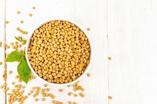 Soy beans in a bowl and on a table, green leaf on wooden board background from above