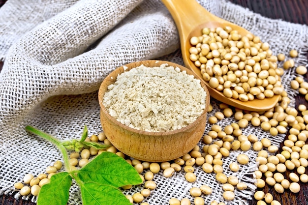 soy flour in bowl, soybeans in spoon on sackcloth, green soya leaf on background of wooden board