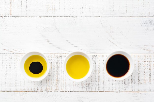 Soy sauce, olive oil and balsamic sauce in ceramic bowls on white wooden surface. Top view.