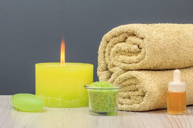 Spa composition with soft terry towel, bottle of aromatic oil, bowl with sea salt, soap and burning candle on gray background.