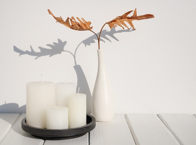 Спа-композиция с белыми свечами и сухим листом филодендрона в современной керамической белой вазе на деревянном столе, интерьер комнаты в земных тонах, длинная тень