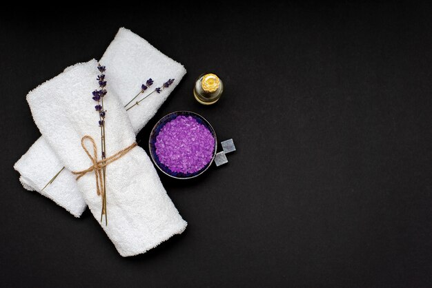 Spa concept. Lavender salt for a relaxing bath, aroma oil, white towels and dry lavender flowers on a black background. Aromatherapy Flat lay.