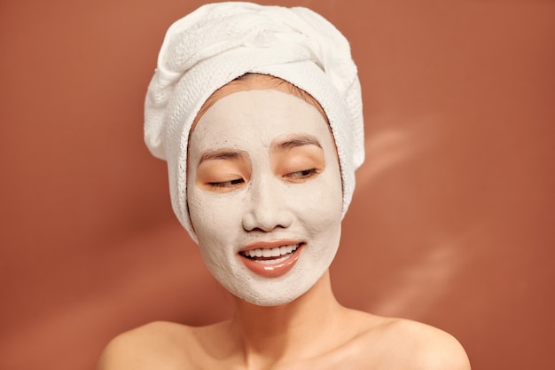 Spa girl with pleased facial expression, applies clay mask on face, gets beauty treatments, wears white soft towel on head