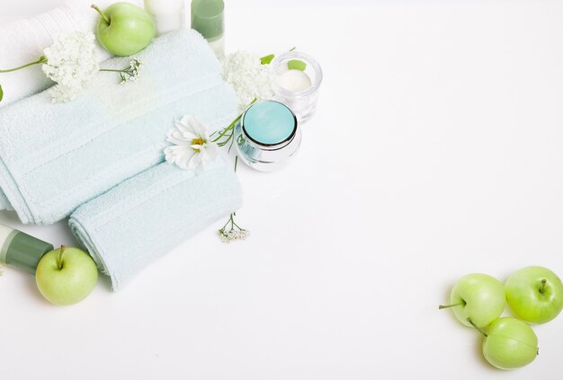 Spa setting on white table. Green apples, towels, cosmos flowers and hydrangeas, cosmetics, candles. Spa resort therapy composition, spa summer concept. Top view