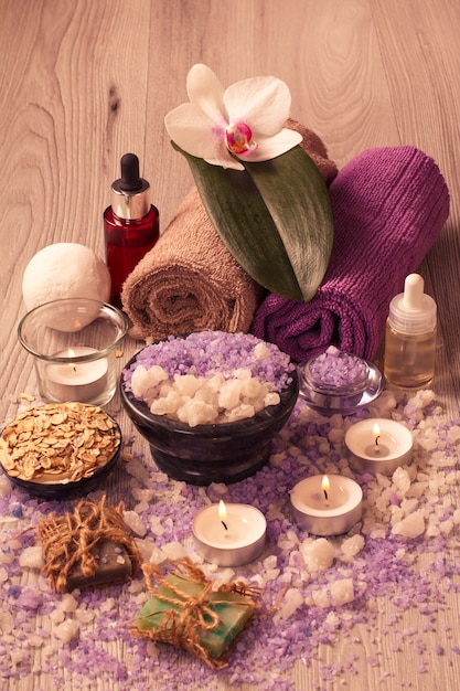 Spa setting with orchid flower, bowl with sea salt, bottles with aromatic oil, soap, scrub, candles and towels on wooden board