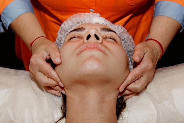스파 전문가는 의료용 모자 미용 치료를 받고 있는 어린 소녀에게 얼굴 마사지를 하고 있습니다.