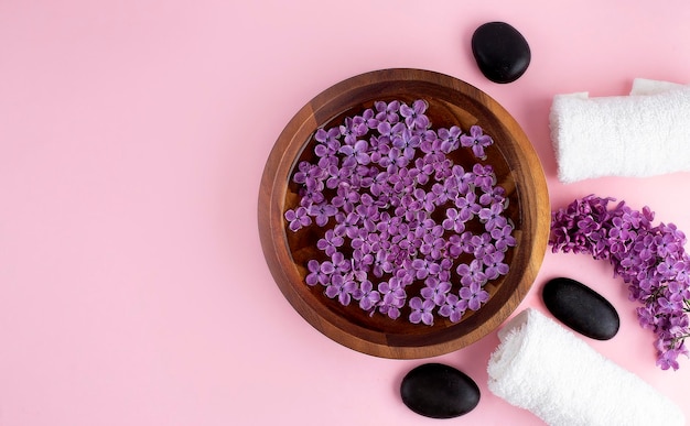 Spa and wellness concept with lilac flowers in water in a wooden bowl and towel aromatherapy and black stones top view flat lay pink background copy space