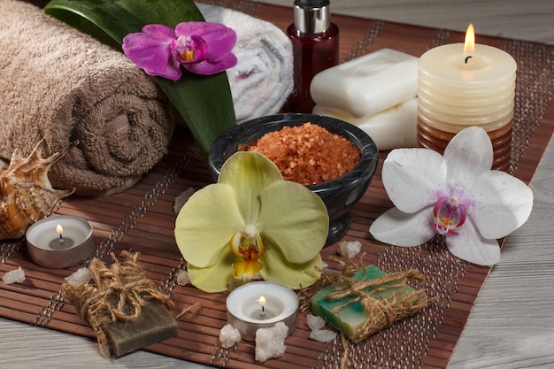 Spa and wellness setting with orchid flowers, bowl with sea salt, seashell, bottle with aromatic oil, soap, candles and towels on bamboo napkin