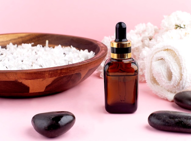 Spa and wellness with perfumed white lilac flowers water in wooden bowl and terry towel and bottle aromatherapy and black stones top view flat lay pink background