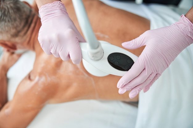 Spa worker wearing gloves and holding massage apparatus to a man back covered in lotion