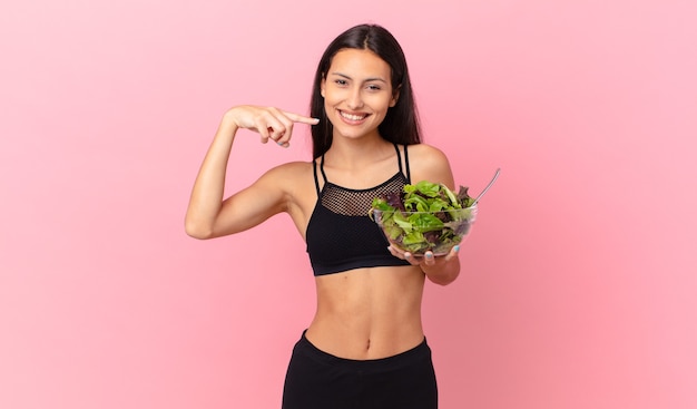 Spaanse fitnessvrouw die zelfverzekerd glimlacht, wijst naar haar eigen brede glimlach en een salade vasthoudt