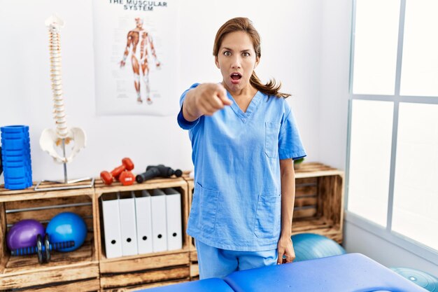 Foto spaanse fysiotherapeut van middelbare leeftijd die werkt bij pijnherstelkliniek en ontevreden en gefrustreerd naar de camera wijst, boos en woedend op jou