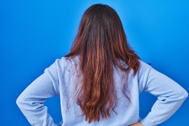 Spaanse jonge vrouw die zich over een blauwe achtergrond bevindt en achteruit kijkt en wegkijkt met de armen op het lichaam