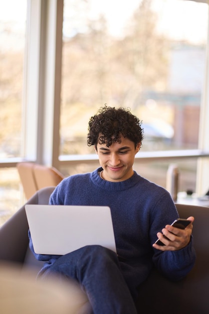 Spaanse jongeman die mobiele telefoon gebruikt terwijl hij op afstand werkt op een laptop in het kantoorinterieur