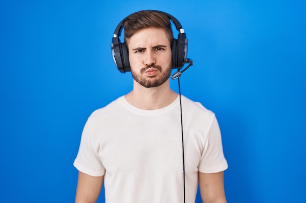 Spaanse man met baard die naar muziek luistert met een koptelefoon die wangen pufft met een grappig gezicht. mond opgeblazen met lucht, gekke uitdrukking.