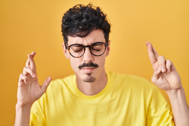 Spaanse man met bril staande over gele achtergrond gebaren vinger gekruist lachend met hoop en gesloten ogen. geluk en bijgelovig concept.