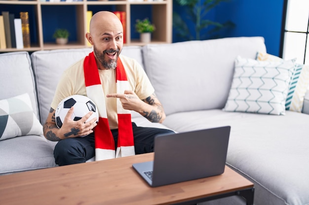 Spaanse man met tatoeages kijken voetbalwedstrijd hooligan bal vasthouden op de laptop glimlachend gelukkig wijzend met hand en vinger