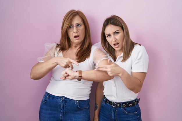 Spaanse moeder en dochter dragen casual wit t-shirt over roze achtergrond in haast wijzend om tijd te kijken ongeduld overstuur en boos voor vertraging deadline