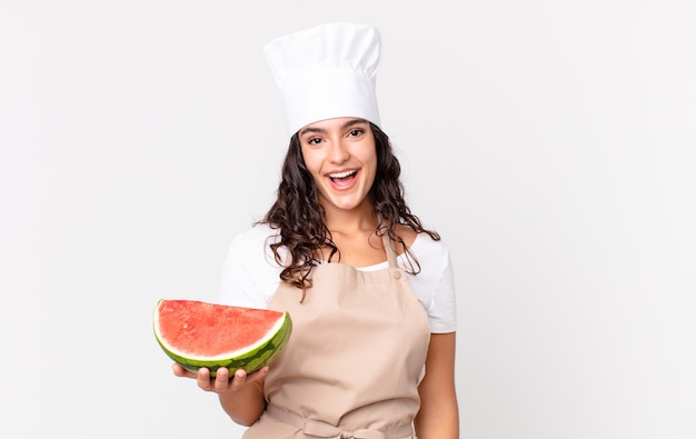 Spaanse mooie chef-kokvrouw die gelukkig en aangenaam verrast kijkt en een watermeloen vasthoudt