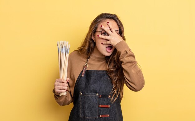 Spaanse mooie vrouw die geschokt, bang of doodsbang kijkt en haar gezicht bedekt met de hand. artiest met borstels concept