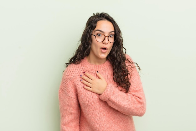 Spaanse mooie vrouw die geschokt en verrast is en glimlacht en haar hand ter harte neemt, blij om degene te zijn of dankbaarheid te tonen