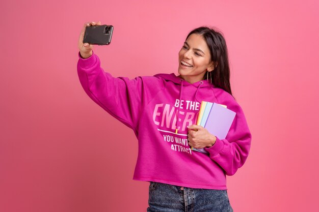 Spaanse mooie vrouw in roze hoodie glimlachend holding notebooks en het gebruik van smartphone poseren op roze geïsoleerd, selfie nemen op camera