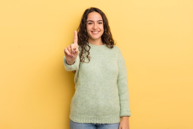 Spaanse mooie vrouw lacht en ziet er vriendelijk uit en toont nummer één of eerste met hand vooruit aftellend