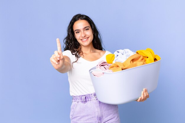 Spaanse mooie vrouw lacht en ziet er vriendelijk uit, toont nummer één en houdt een wasmand vast