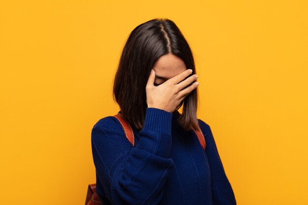 Spaanse vrouw die gestrest, beschaamd of boos kijkt, met hoofdpijn, gezicht bedekt met hand