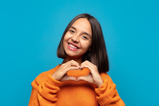 Spaanse vrouw die lacht en zich gelukkig, schattig, romantisch en verliefd voelt, hartvorm maakt met beide handen