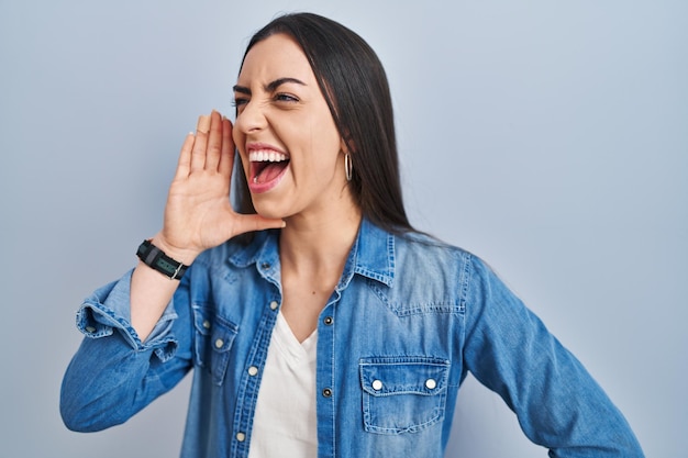 Spaanse vrouw die over een blauwe achtergrond staat te schreeuwen en luid naar de andere kant te schreeuwen met de hand op de mond. communicatieconcept.