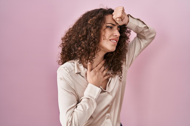Spaanse vrouw met krullend haar dat over een roze achtergrond staat en het voorhoofd aanraakt voor ziekte en koorts, griep en verkoudheid, virusziek