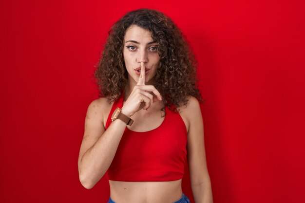 Spaanse vrouw met krullend haar die over een rode achtergrond staat en vraagt om stil te zijn met de vinger op de lippen. stilte en geheim concept.