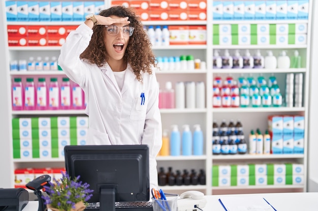 Spaanse vrouw met krullend haar werken bij apotheekdrogisterij erg blij en glimlachend ver weg kijkend met hand boven hoofd zoeken concept