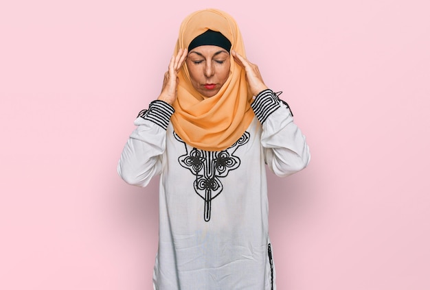 Spaanse vrouw van middelbare leeftijd die traditionele islamitische hijab-sjaal draagt en lijdt aan hoofdpijn, wanhopig en gestrest vanwege pijn en migraine. handen op het hoofd.