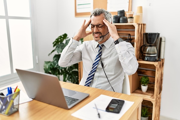 Spaanse zakenman van middelbare leeftijd die op kantoor werkt en een headset draagt met de hand op het hoofd vanwege pijn in het hoofd vanwege stress. last van migraine.