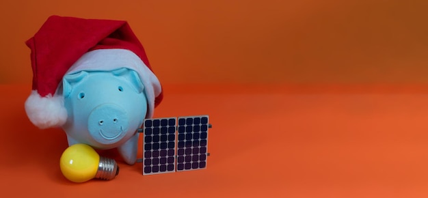 Spaarvarken munten met kerstman kerstmuts, fotovoltaïsch zonnepaneel en lamp geïsoleerd op een oranje achtergrond. Concept afbeelding.