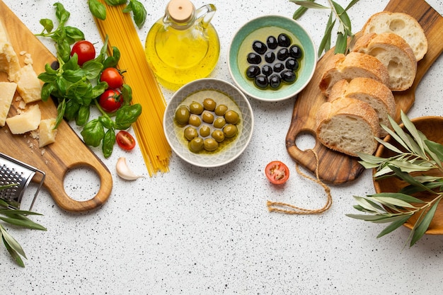 Spaghetti, cheese parmesan, ripe olives, ciabatta, olive tree sprigs and snacks on white rustic background. Ingredients and appetizers for Italian pasta or mediterranean meal, top view, copy space