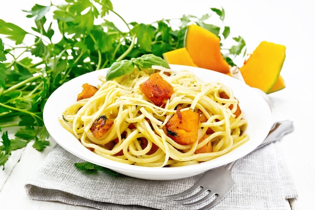 Spaghetti met pompoen, ui en knoflook in plaat op een handdoek, basilicum, peterselie en een vork op houten raadsachtergrond