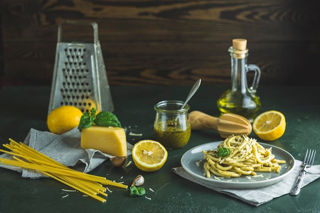 Паста спагетти букатини с соусом песто и пармезаном. Итальянская традиционная паста перчателли с соусом песто Дженовезе в сером блюде, подается с ингредиентами на темно-зеленом бетонном столе.
