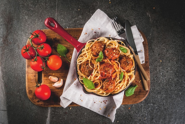 Spaghetti pasta met gehaktballetjes, basilicum tomatensaus in rode gietijzeren pan, op zwarte stenen tafel met snijplank